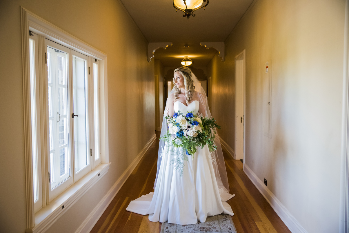 A bride standing in a hallway holding a bouquet, looking out the window, captured by Denver wedding photographer, Casey Van Horn.