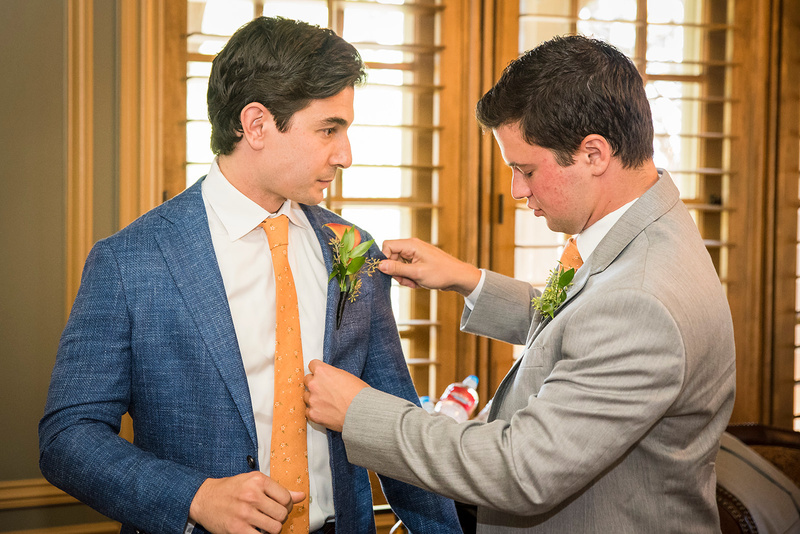 A groom and groomsman in formal attire are fixing each other's ties.