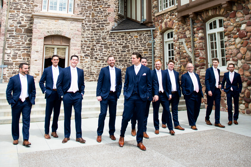 A groom and groomsmen in navy blue suits standing in front of a stone building.